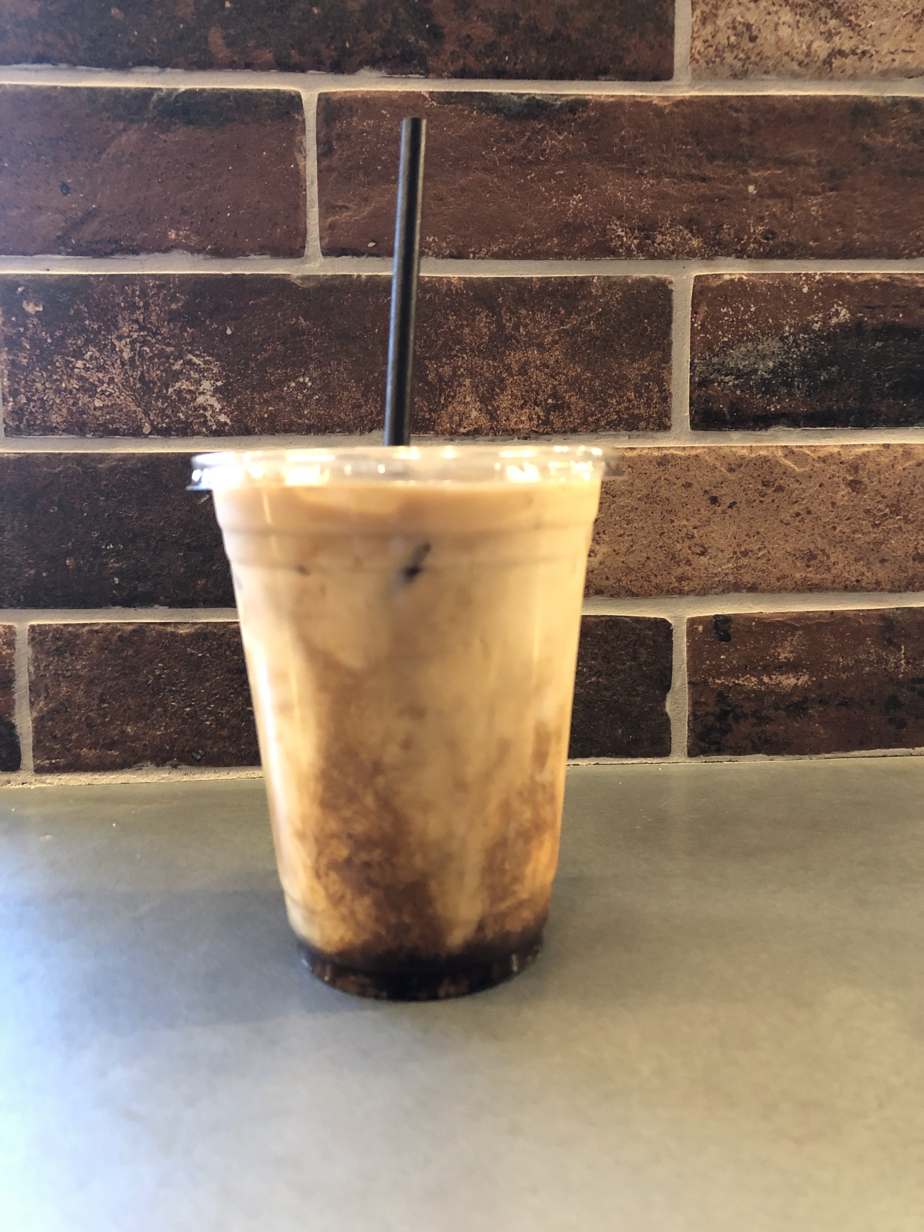 An iced coffee in a clear plastic cup site on a metal countertop in front of a brick wall. The cup has a black straw in it.